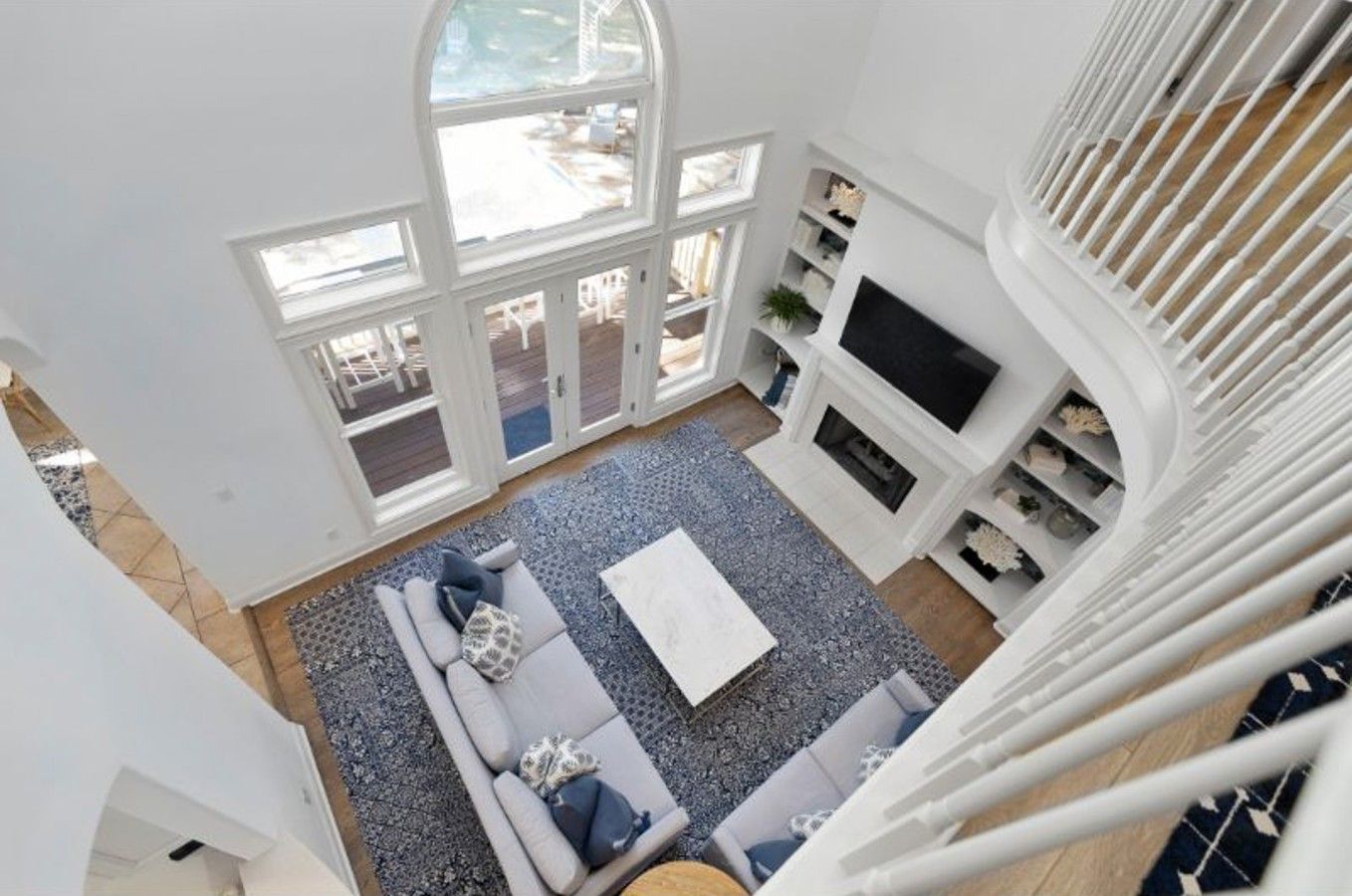 A high-ceiling living room with a large window, a sofa set, a flat-screen TV, and a fireplace.