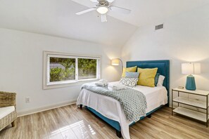Un dormitorio con cama, ventilador de techo, mesita de noche con lámpara y ventana con persianas.