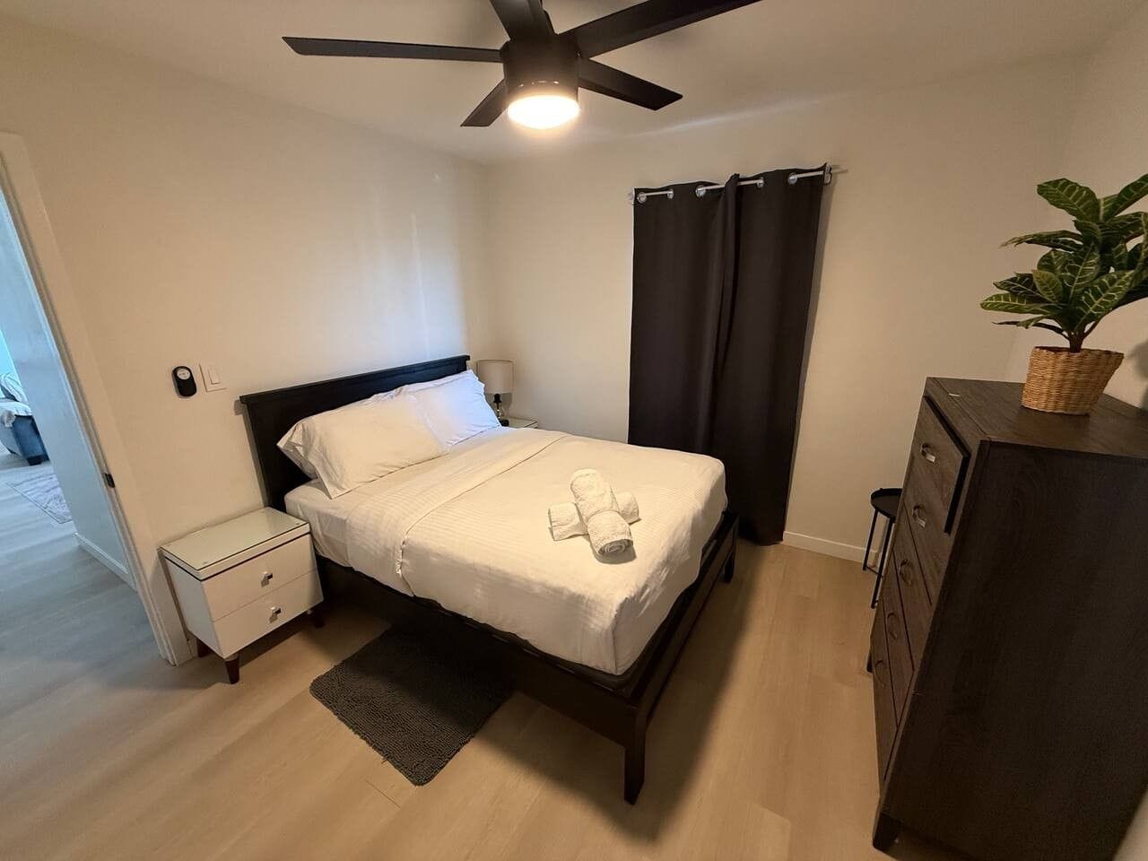 A neatly made bed with a white comforter and a rolled towel, a small bedside table, a dark-colored dresser, a ceiling fan, and a potted plant on the dresser.