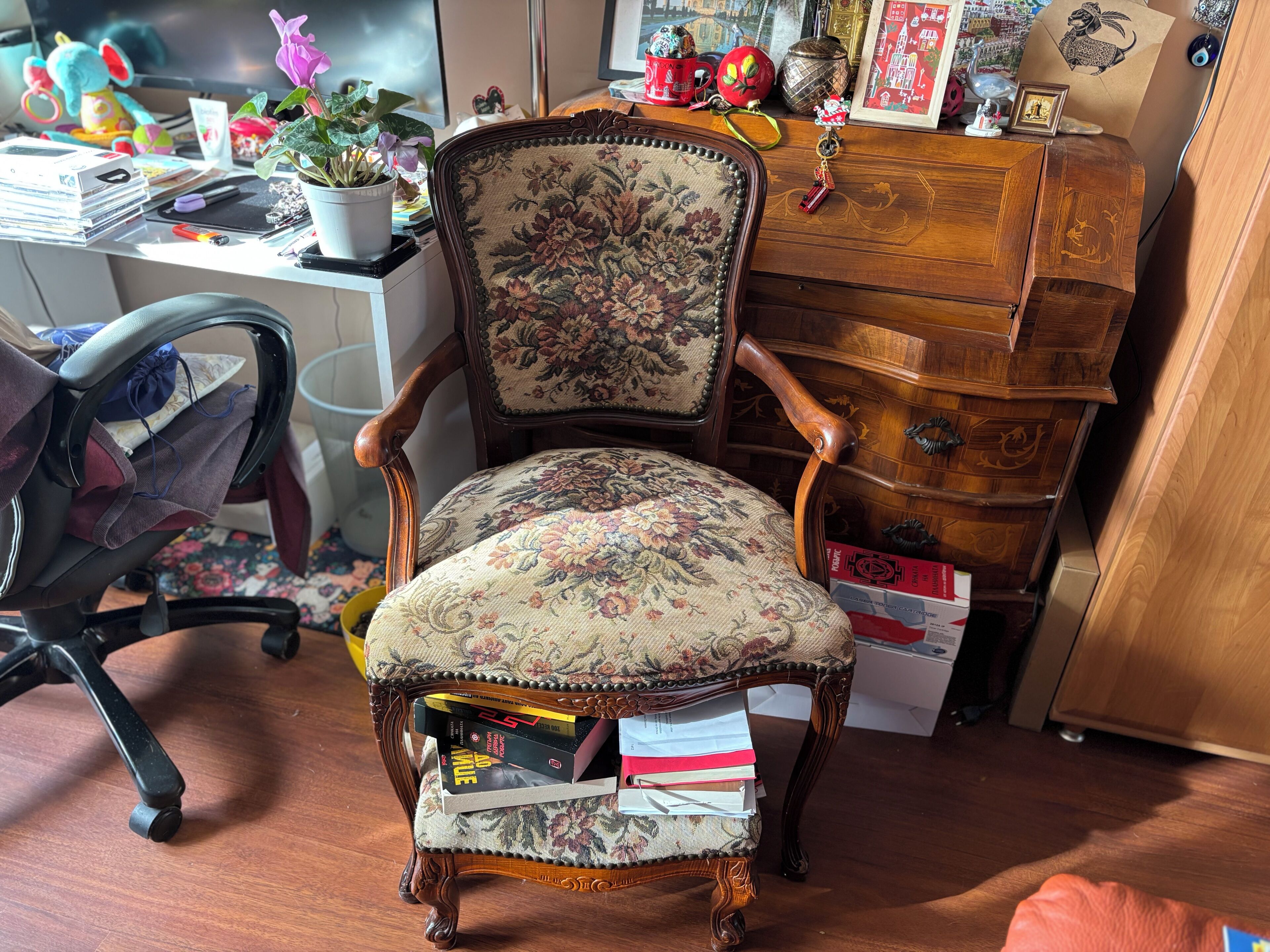 A vintage-style chair with floral upholstery, a wooden desk with various items, and a wooden cabinet with drawers.