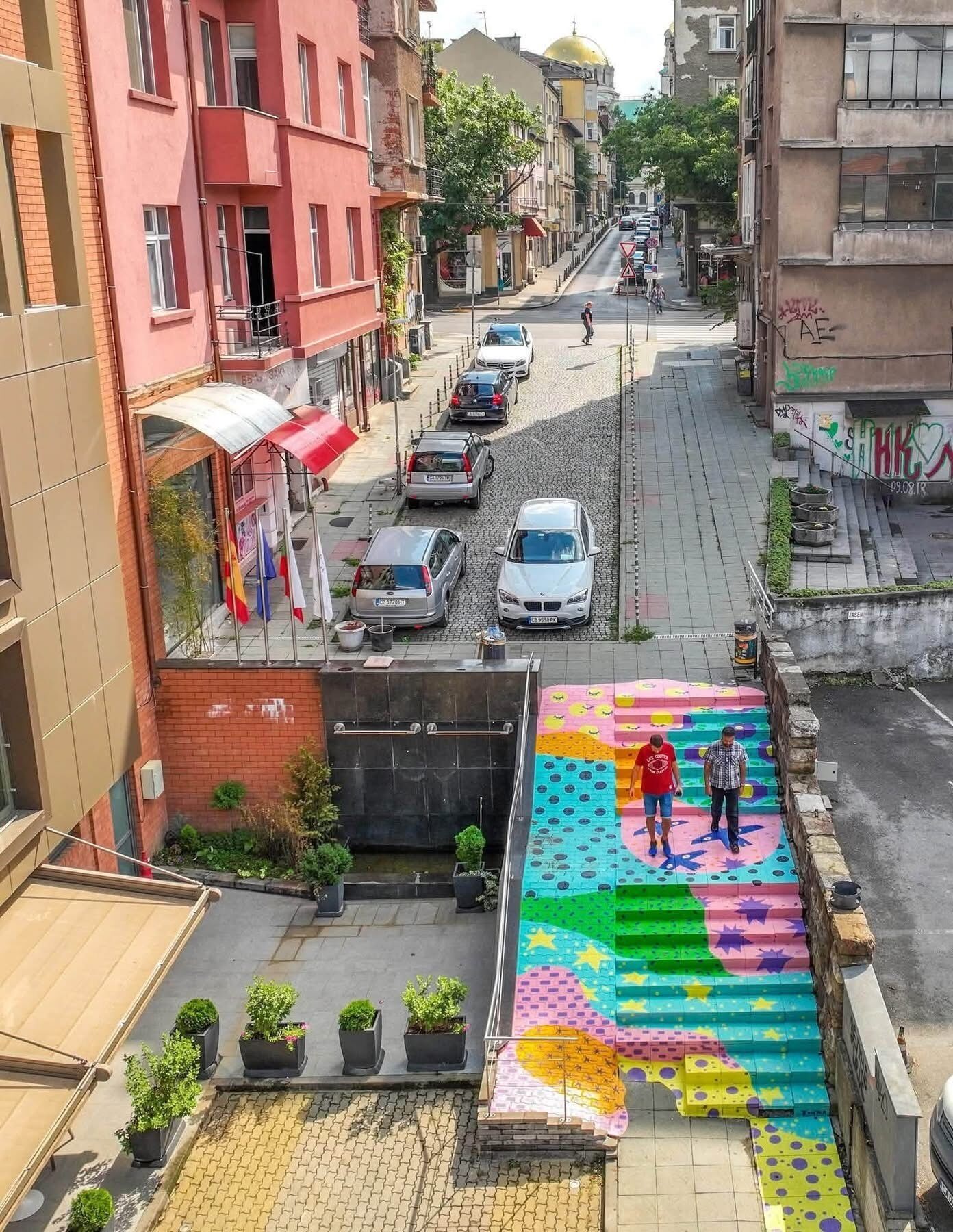 A street with parked cars, a pedestrian crossing, and buildings with balconies.