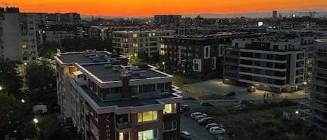 A multi-story residential building with a flat roof, surrounded by other buildings, and a parking area with several cars.