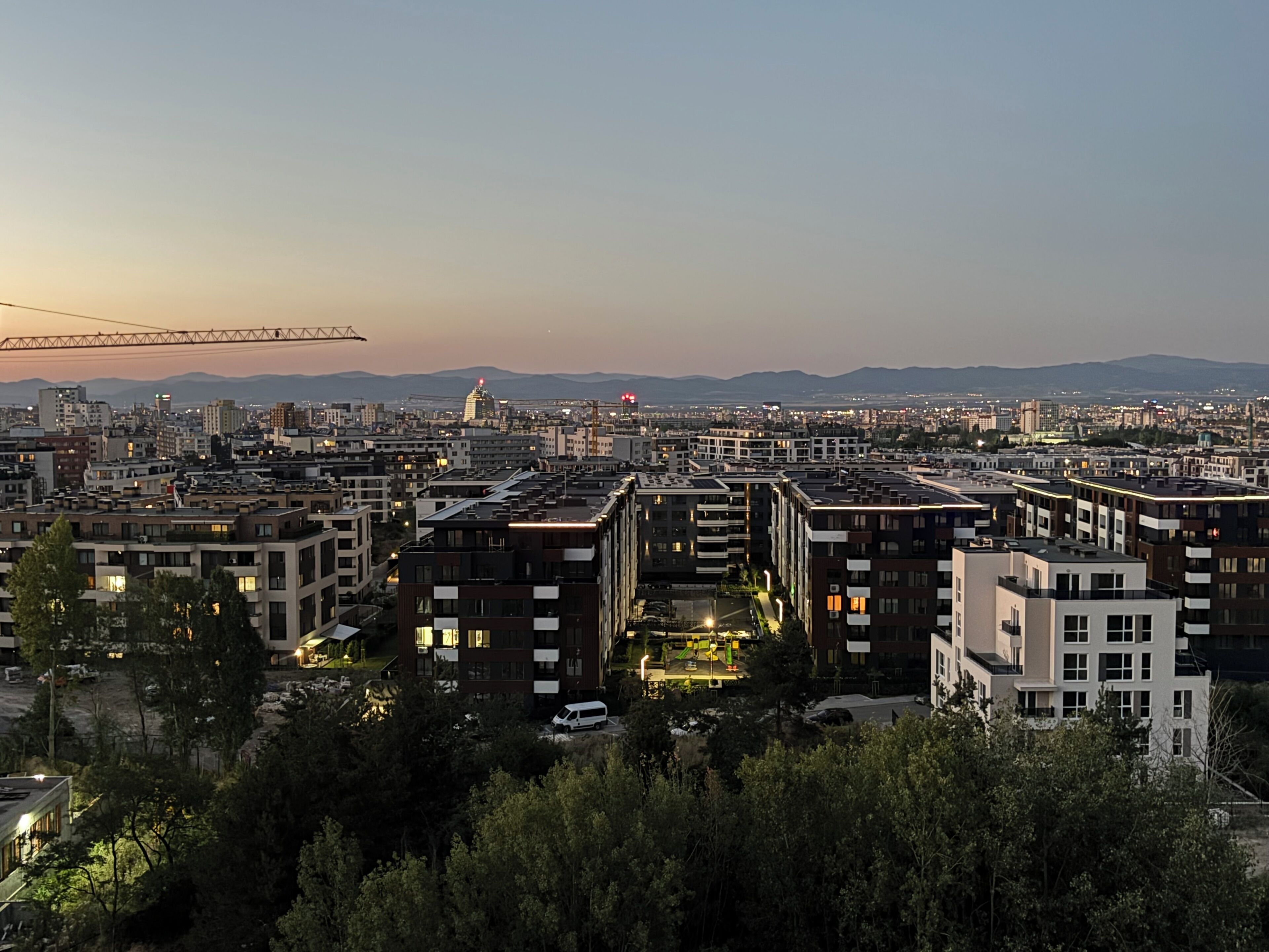 A cityscape at dusk with modern buildings, a construction crane, and distant mountains.