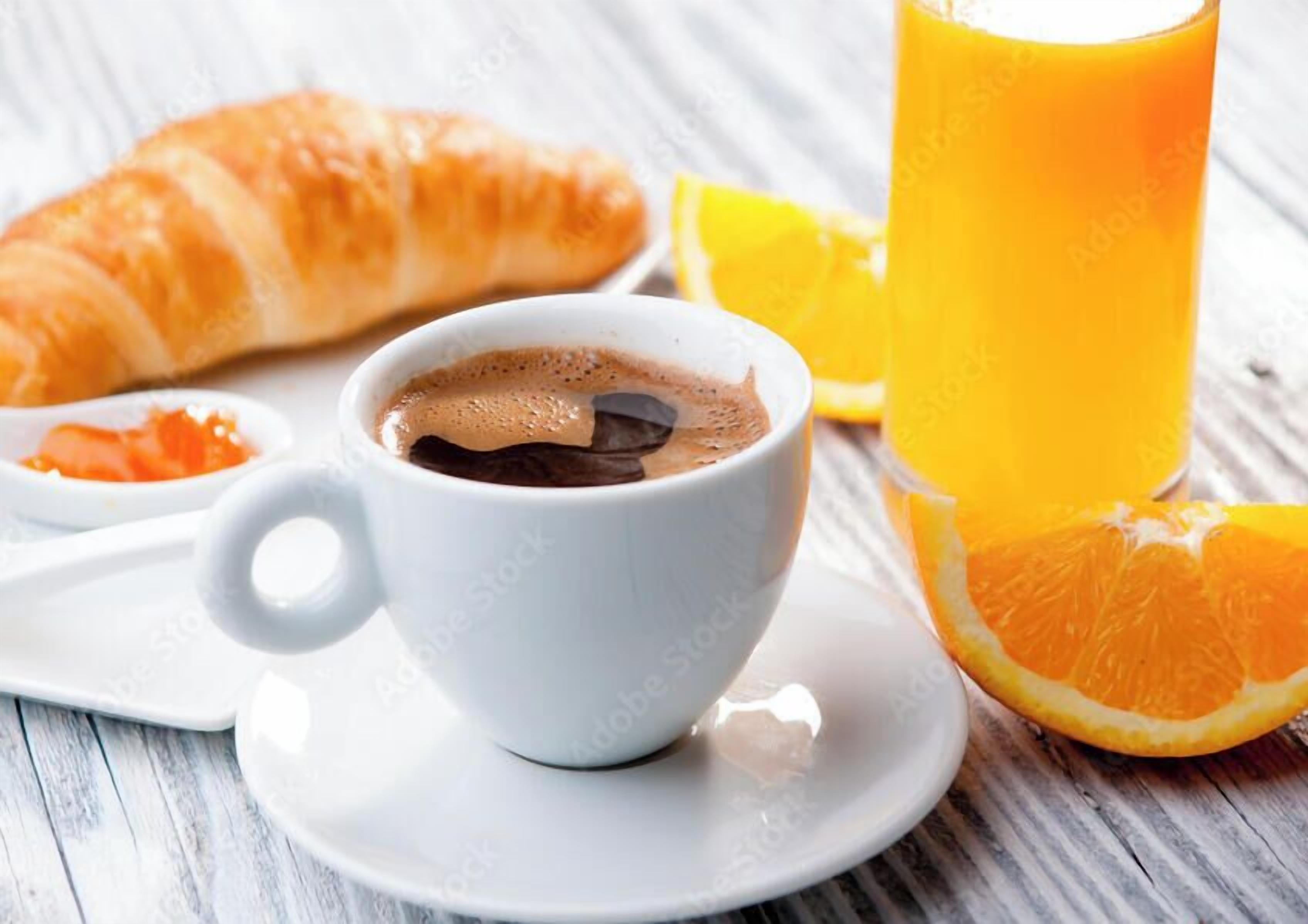 Una taza de café blanca con un platito, un croissant y un vaso de zumo de naranja sobre una mesa de madera.