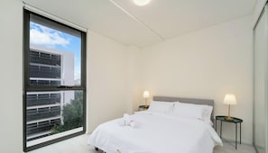 Une chambre d’hôtel avec une grande fenêtre, un lit avec une literie blanche, deux tables de chevet avec des lampes et une vue sur un bâtiment à l’extérieur.