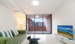 A modern living room with a sofa, a coffee table, a flat-screen TV, and a sliding glass door leading to a balcony with a cityscape view. - MetaWiseBnB 2BR Near Airport Parking Sleeps 6 (Mascot)