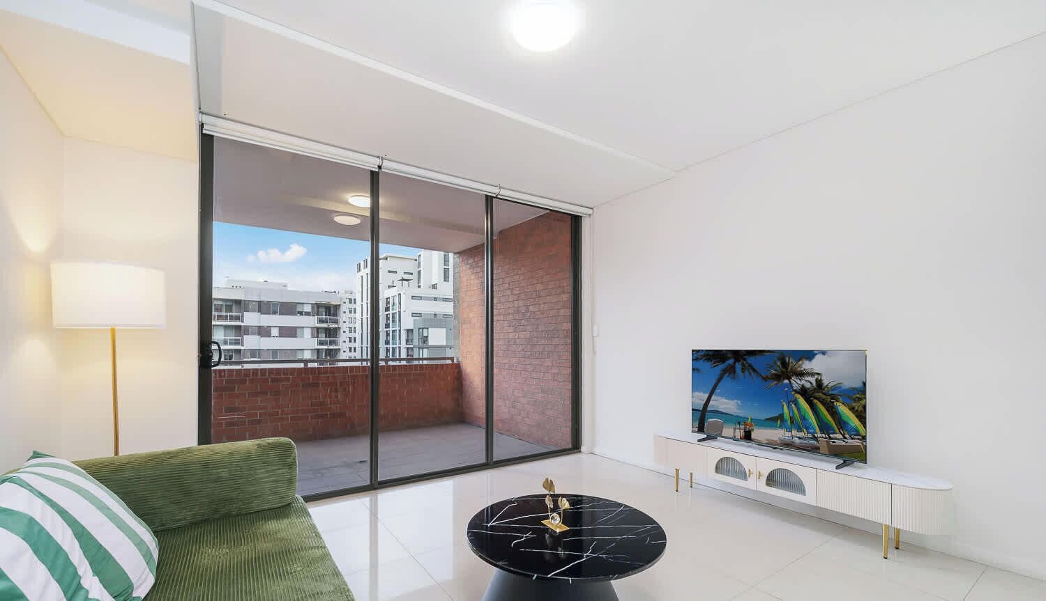 Uma sala de estar moderna com um sofá, uma mesa de centro, uma TV sobre um móvel branco e uma janela grande com vista para a cidade.