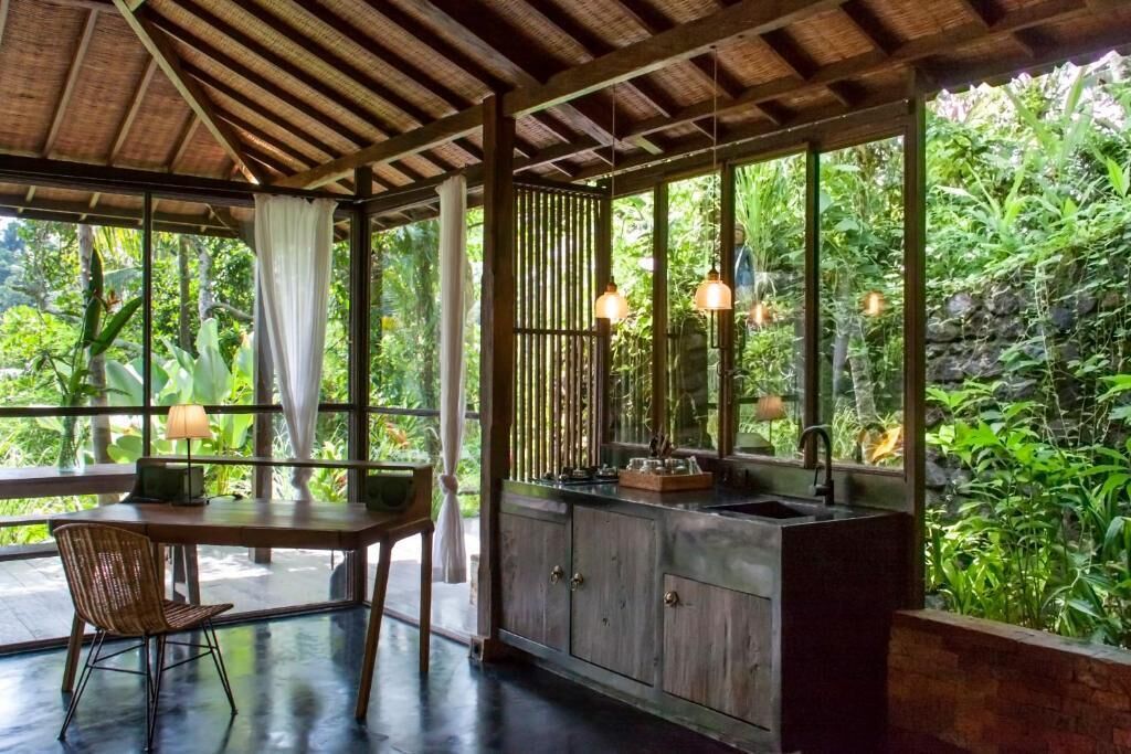 A room with a wooden ceiling, a dining table with chairs, a kitchen area with a sink, and large windows overlooking greenery.