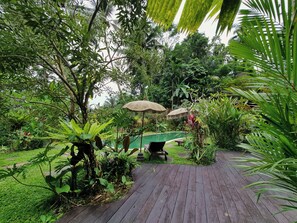 A wooden deck leading to a pool area surrounded by lush greenery, with two outdoor umbrellas providing shade. - Villa Mahadevi (Gianyar)