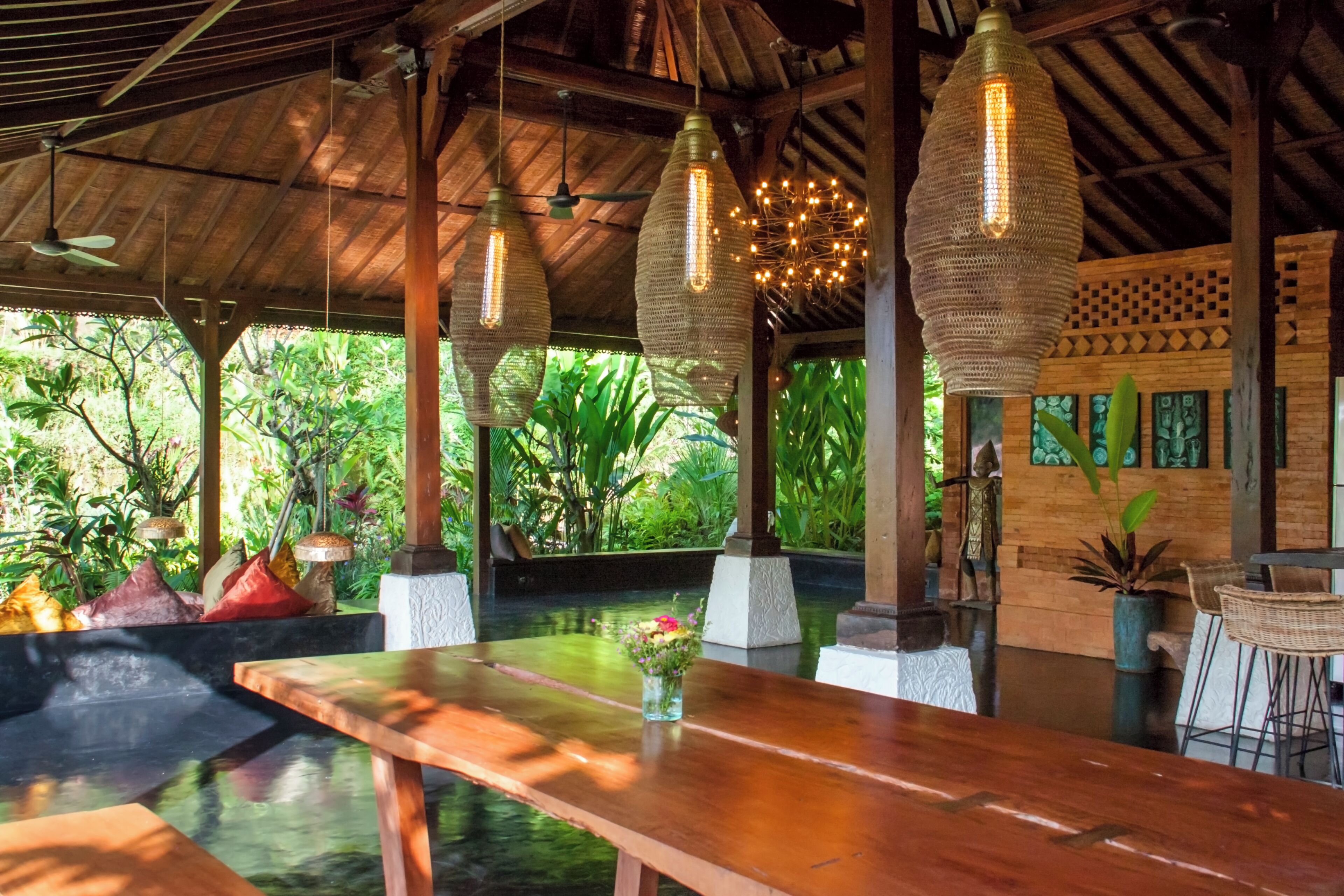 A wooden table with a vase of flowers, hanging woven light fixtures, and a view of lush greenery outside.