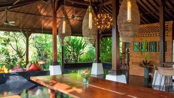 Una mesa de madera con un jarrón de flores, lámparas colgantes tejidas y vistas de vegetación exuberante al exterior.