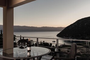 Une table ronde en verre, avec deux verres à vin et une bouteille, une chaise en osier et une vue sur un lac où l’on aperçoit des bateaux et des collines.