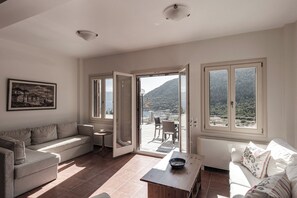 En stue med sofa, sofabord og spiseområde med stole og bord. En dør fører ud til en balkon med udsigt til et bjerg.