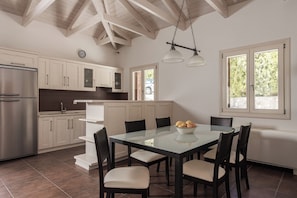 A modern kitchen with a central island, stainless steel appliances, and a dining area with a glass table and chairs. - Villa Idylle | Ithaca Greece (Ithaki)
