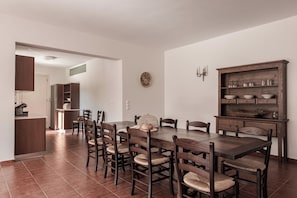 A dining area with a large wooden table and chairs, a kitchen area with cabinets, and a wall-mounted shelf with glassware. - Villa Idylle | Ithaca Greece (Ithaki)