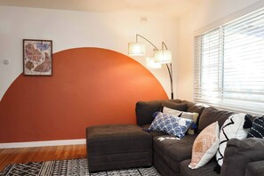 Ein Wohnzimmer mit einer Couch, einem Teppich, einer Wand mit einer großen orangefarbenen Form und einem Fenster mit Jalousien.