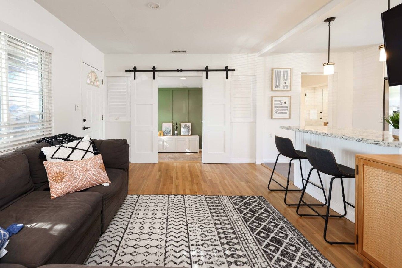 A modern living room with a brown sofa, patterned rug, and a kitchen area with bar stools and a countertop.