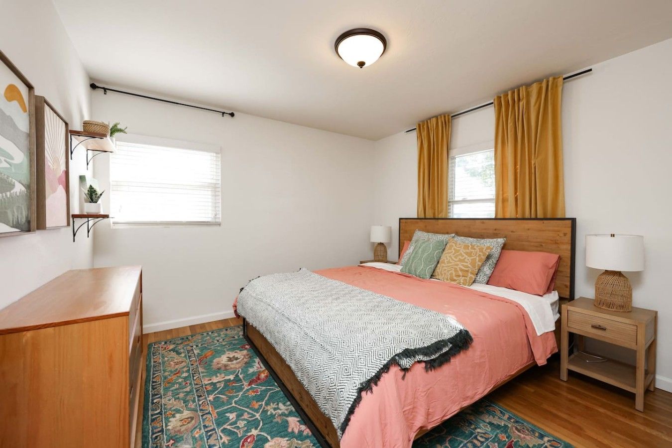 A bedroom with a bed, bedside tables, a nightstand, a window with curtains, a wall-mounted shelf with plants, and a patterned rug.