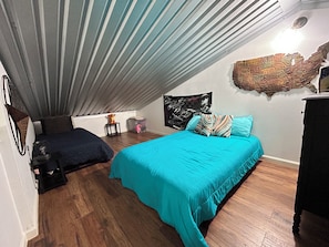 A bedroom with a sloped ceiling, a bed with a teal bedspread, a small table with a lamp, and a wall-mounted tapestry.