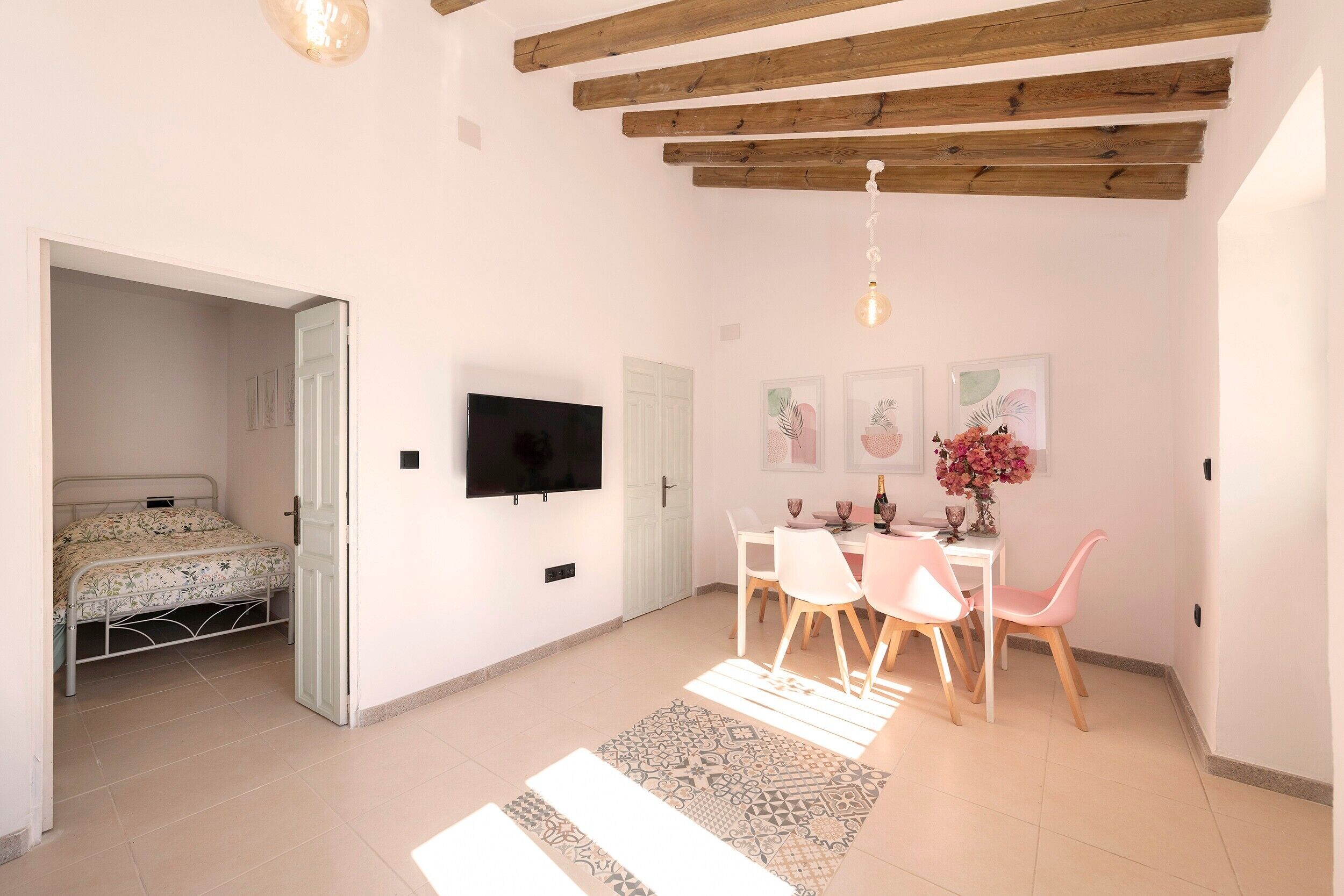 Een kamer met een bed, een aan de muur bevestigde televisie, een eettafel met stoelen en een plafond met zichtbare houten balken.