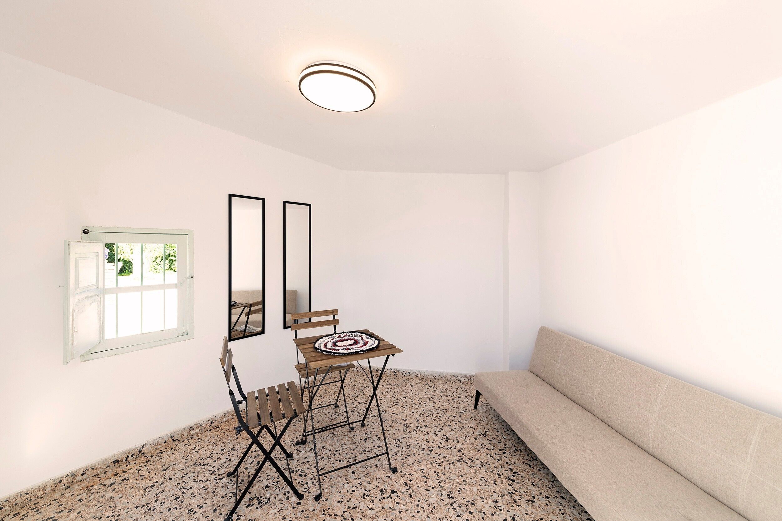 A minimalist living room with a sofa, a small table, two chairs, a mirror, and a window with a view of greenery.