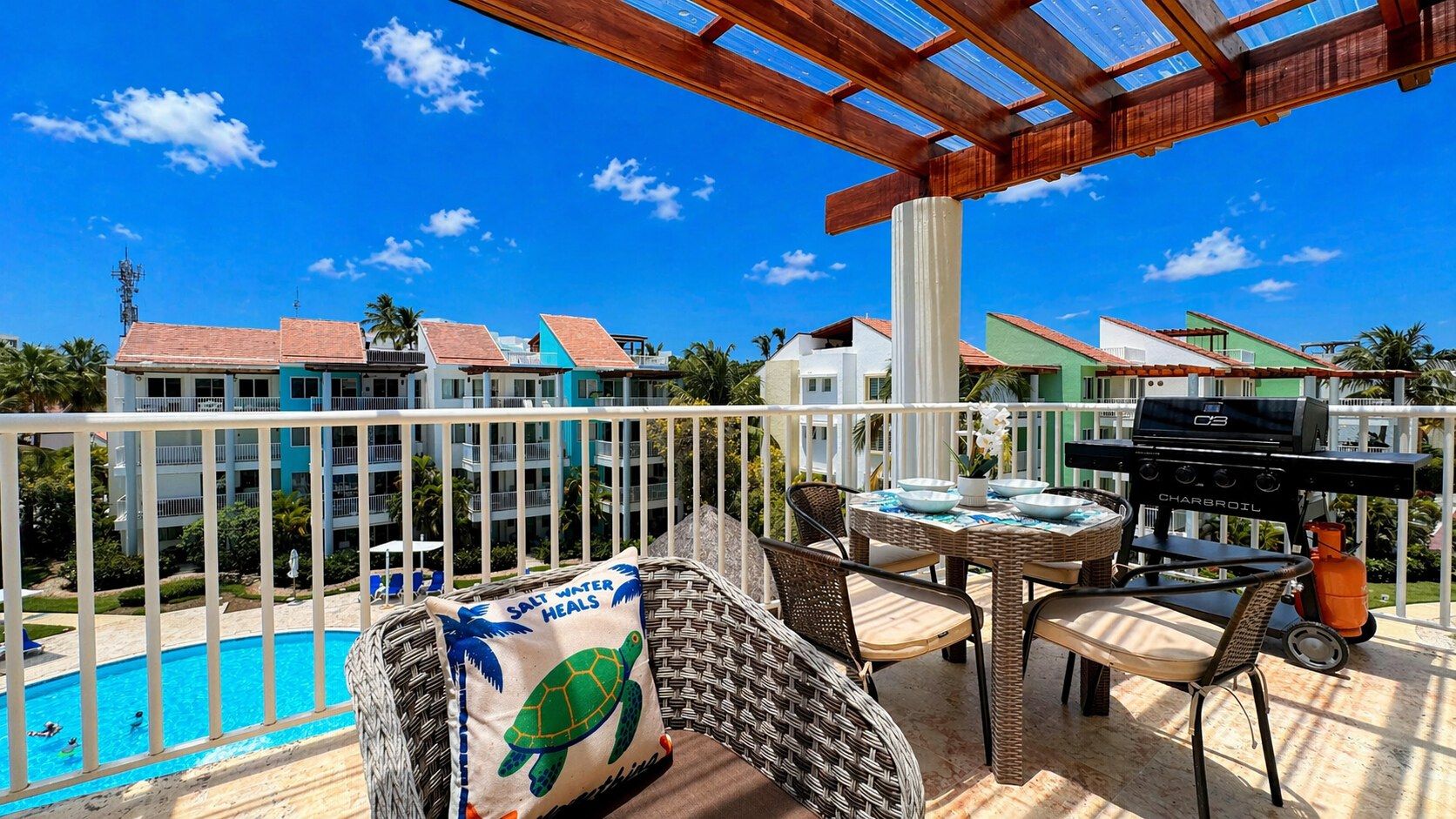 A balcony with wicker furniture, a barbecue grill, and a view of a swimming pool and buildings.