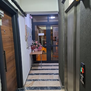 Un pasillo con una puerta de madera, una pequeña mesa con un jarrón de flores y un suelo de baldosas.
