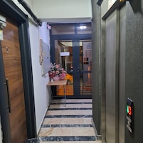 Un pasillo con una puerta de madera, una pequeña mesa con un jarrón de flores y un suelo de baldosas.