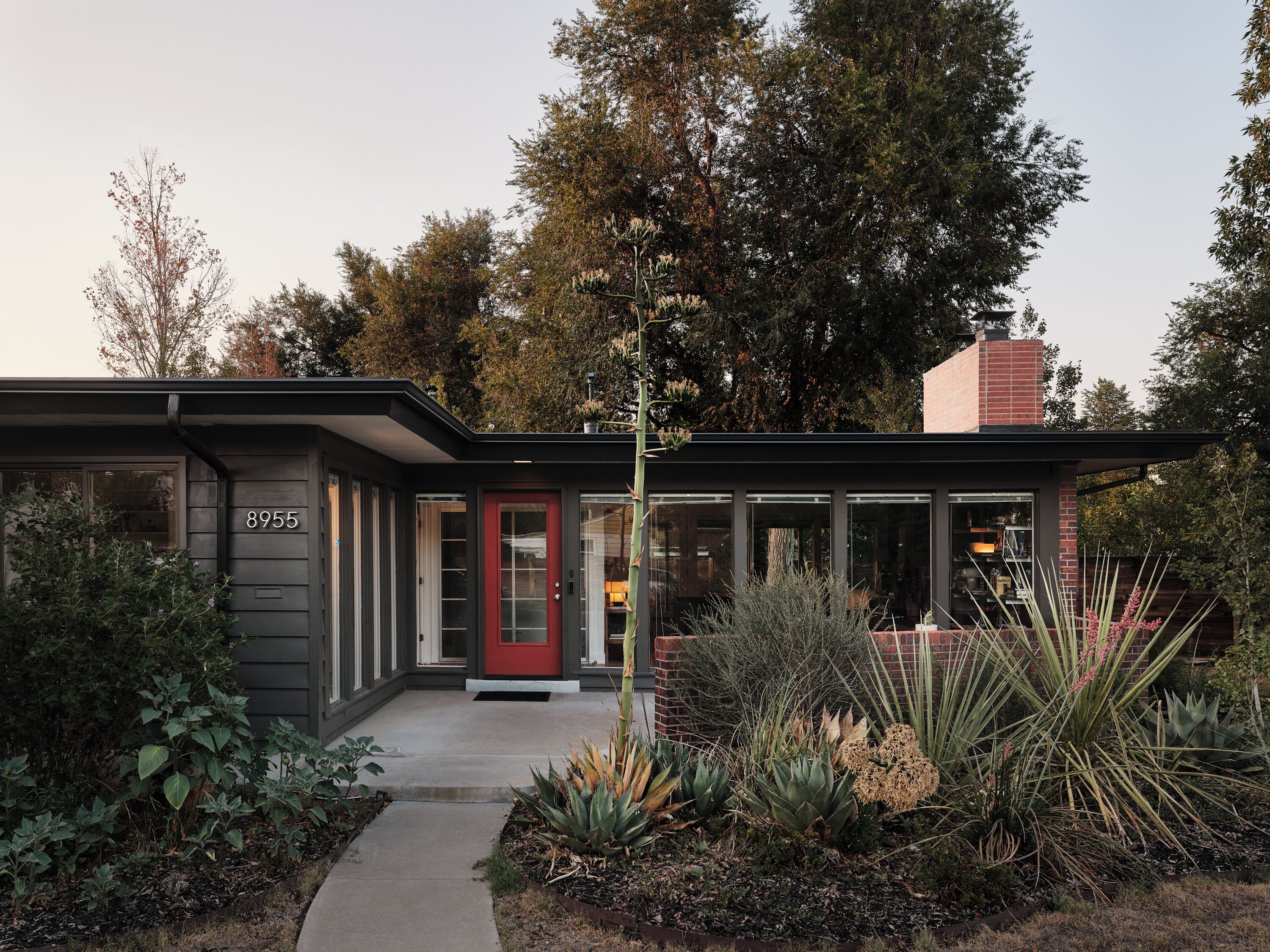 Una casa moderna con puerta roja, número 8955, rodeada por un jardín con diversas plantas.