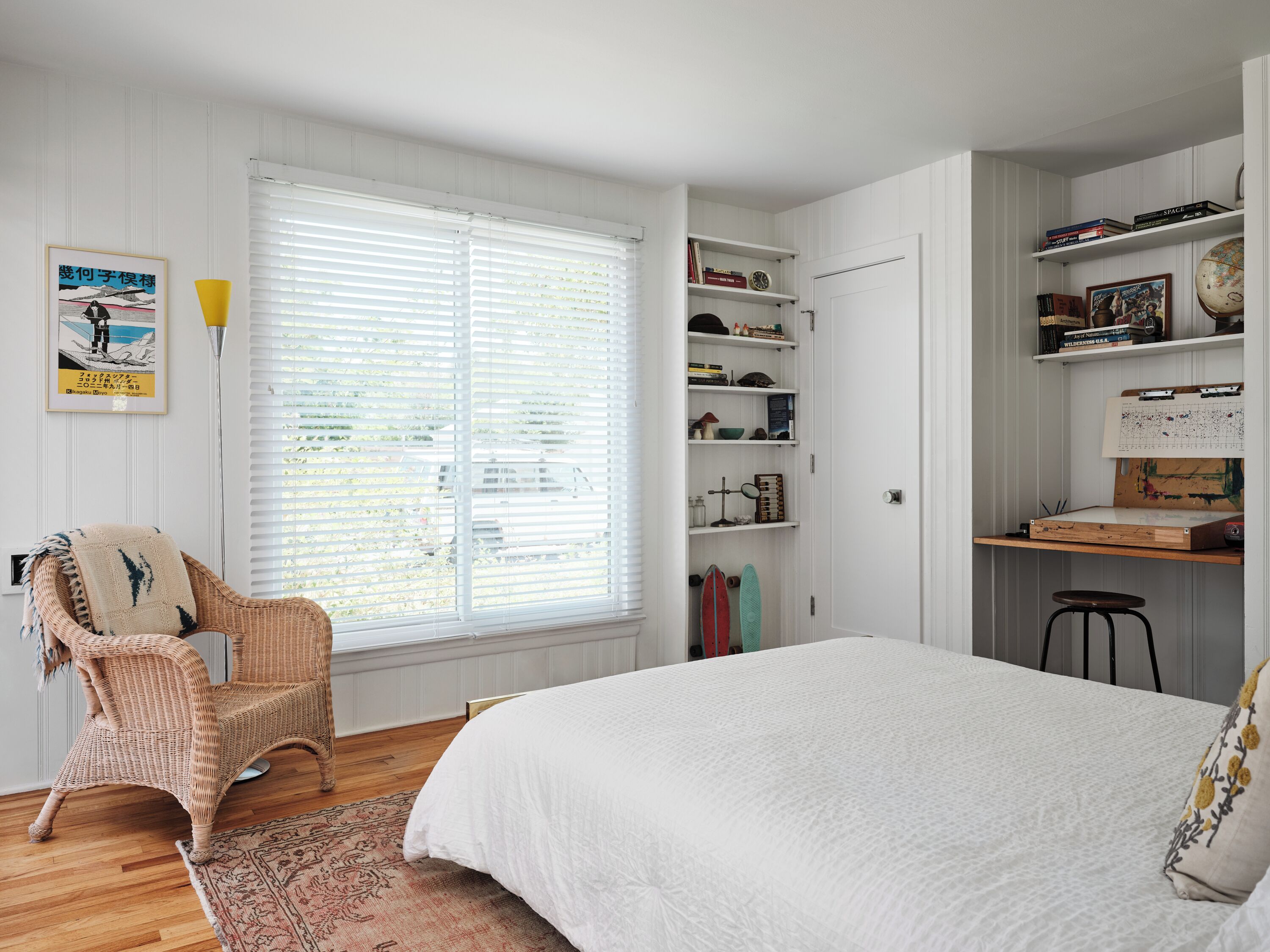 A bedroom with a large bed, a wicker chair, a desk with shelves, and a window with blinds.