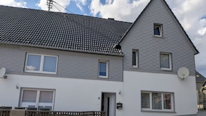 Um edifício residencial de dois andares, com telhado de telhas cinzas, paredes externas brancas e um pequeno alpendre frontal.