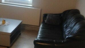 Uma sala de estar com um sofá de couro preto, uma mesa pequena com um objeto amarelo e uma janela com persianas.