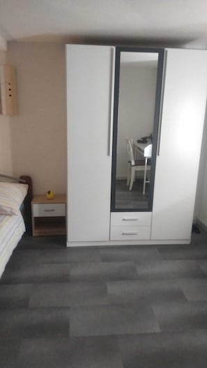 A bedroom with a bed, a wardrobe, a mirror, a small table with a lamp, and a chair.