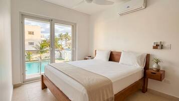 A bedroom with a bed, a bedside table, a nightstand, a ceiling fan, an air conditioning unit, and a view of a swimming pool and palm trees.
