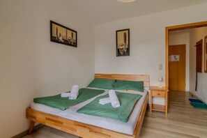 Uma cama de solteiro com estrutura de madeira, roupa de cama verde e duas toalhas brancas. Há uma mesa de cabeceira com uma luminária e uma gavetinha. O quarto tem paredes lisas e piso de madeira.