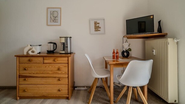 A compact kitchenette with a wooden cabinet, a coffee maker, a kettle, and a microwave on a shelf. There are two white chairs and a small table with a vase and two bottles. - Cozy 1-room apartment, sauna, swimming pool, parking (Zwiesel)
