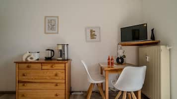 A compact kitchenette with a wooden cabinet, a coffee maker, a kettle, and a microwave on a shelf. There are two white chairs and a small table with a vase and two bottles.