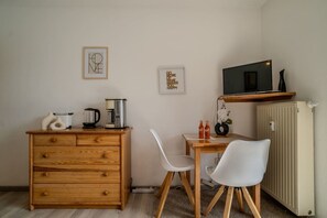 Una kitchenette compacta con un mueble de madera, una cafetera, un hervidor y un microondas sobre una estantería. Hay dos sillas blancas y una mesa pequeña con un florero y dos botellas.