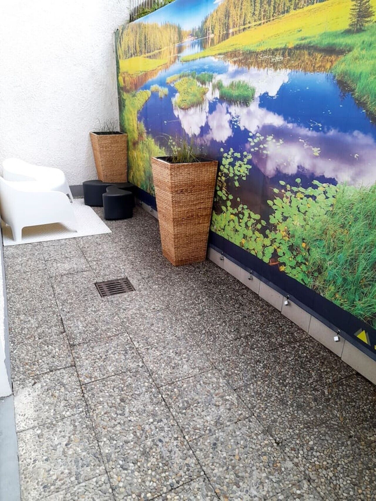 Ein Fliesenboden mit einem großen Gemälde von einem See und Grünflächen, zwei Korbmöbelpflanzgefäßen und einem weißen Stuhl.