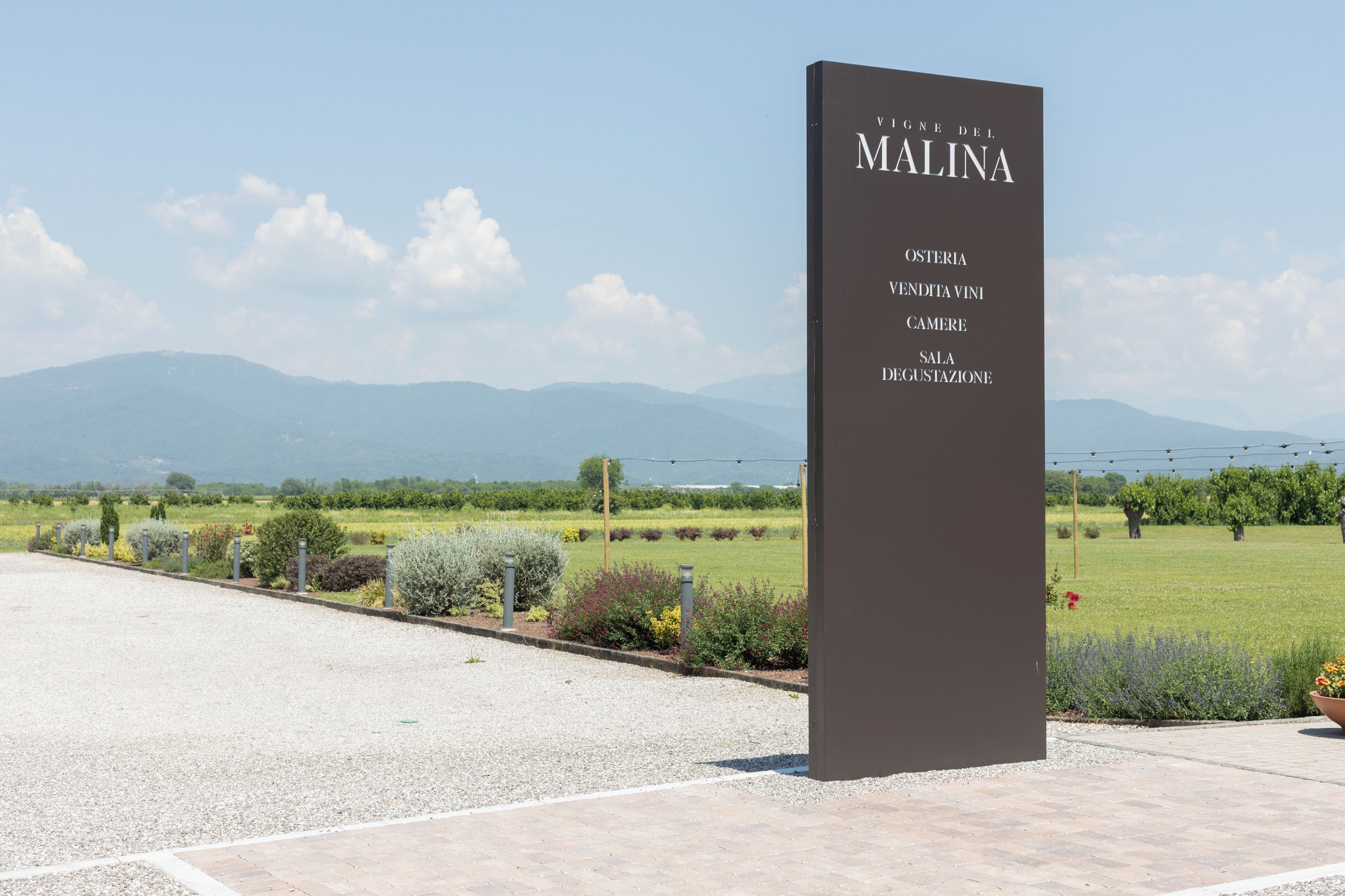 En skylt med texten ”Vigne del Malina” och ”Osteria Vendita Vini Camera Sala Degustazione” placerad på ett väl synligt utomhusområde med utsikt över avlägsna berg.
