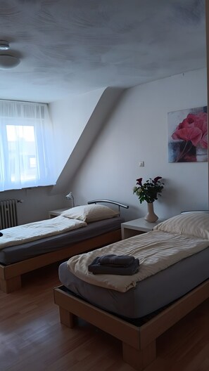 A room with two beds, a radiator, a window with sheer curtains, a bedside lamp, a wall-mounted power outlet, and a painting of flowers. - Apartment 'Haus Abendsonne Top Floor' with Terrace, Garden & Wi-Fi (Koblenz)