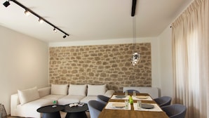 A modern living room with a stone wall, a wooden dining table set for two, and a sofa. - Kamak Villa I (West Mani)