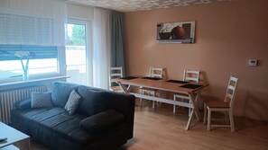 A furnished living room with a sofa, a dining table with chairs, a window with curtains, and a painting on the wall. - Apartment 'Haus Abendsonne Großes Hochparterre' with Private Terrace, Shared Garden and Wi-Fi (Koblenz)
