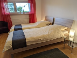 Ein Doppelbett mit Holz-Kopfteil, ein Nachttisch mit Lampe und ein Fenster mit roten Vorhängen.