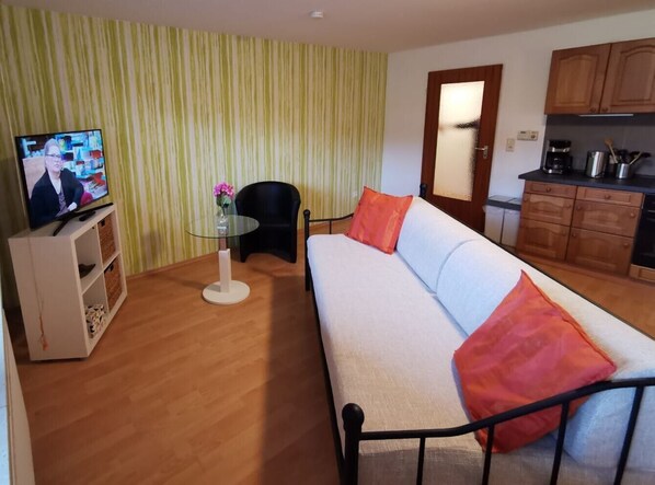 Ein Wohnzimmer mit einer Couch, einem Glastisch, einem Fernseher und einer Küchenzeile mit Schränken.