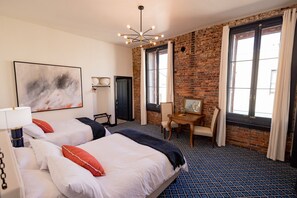 A hotel room with two beds, a desk, a chair, a large window, and a brick wall.
