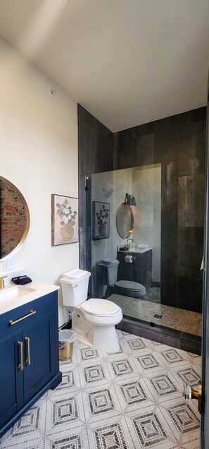 A modern bathroom with a glass shower enclosure, a white toilet, a blue vanity with gold handles, and a large mirror.