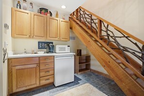 Eine Küche mit Holzschränken, einem weißen Kühlschrank, einer Mikrowelle und einer Kaffeemaschine. Es gibt eine Holztreppe mit Geländer.