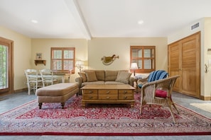 A living room with a sofa, ottoman, and a chair, a wooden cabinet, and a red patterned rug.