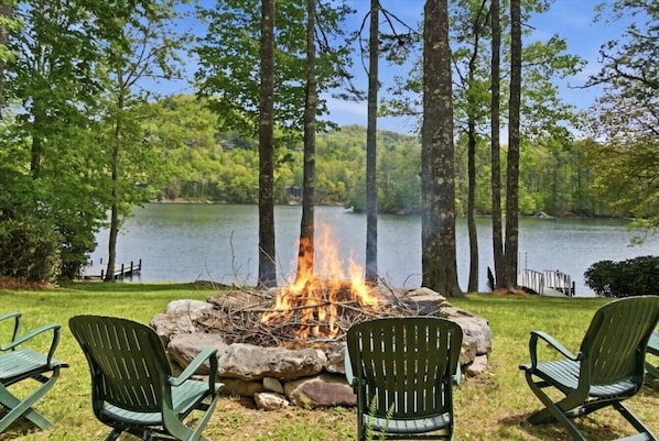 A lakeside campsite with a fire pit surrounded by chairs, a stone wall, and tall trees.